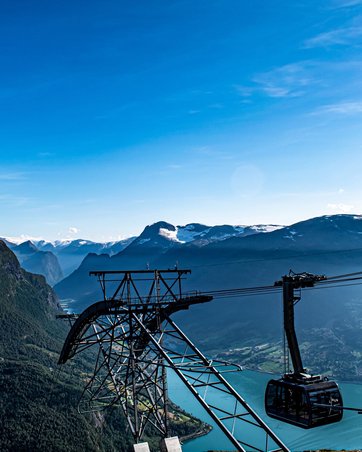 Gondola lift at Mount Hoven in Loen, Norway, overlooking turquoise Nordfjord, steep green mountains and distant snow-covered summits.