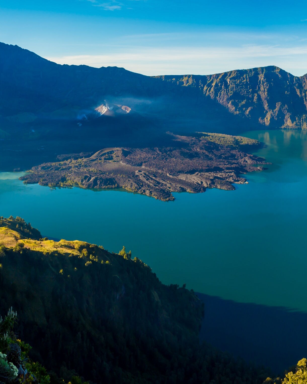 Panoramic view of Mount Rinjani’s crater, featuring a vivid turquoise lake, steep rocky slopes and a small volcanic cone rising from the center, surrounded by lush green ridges under a clear blue sky.