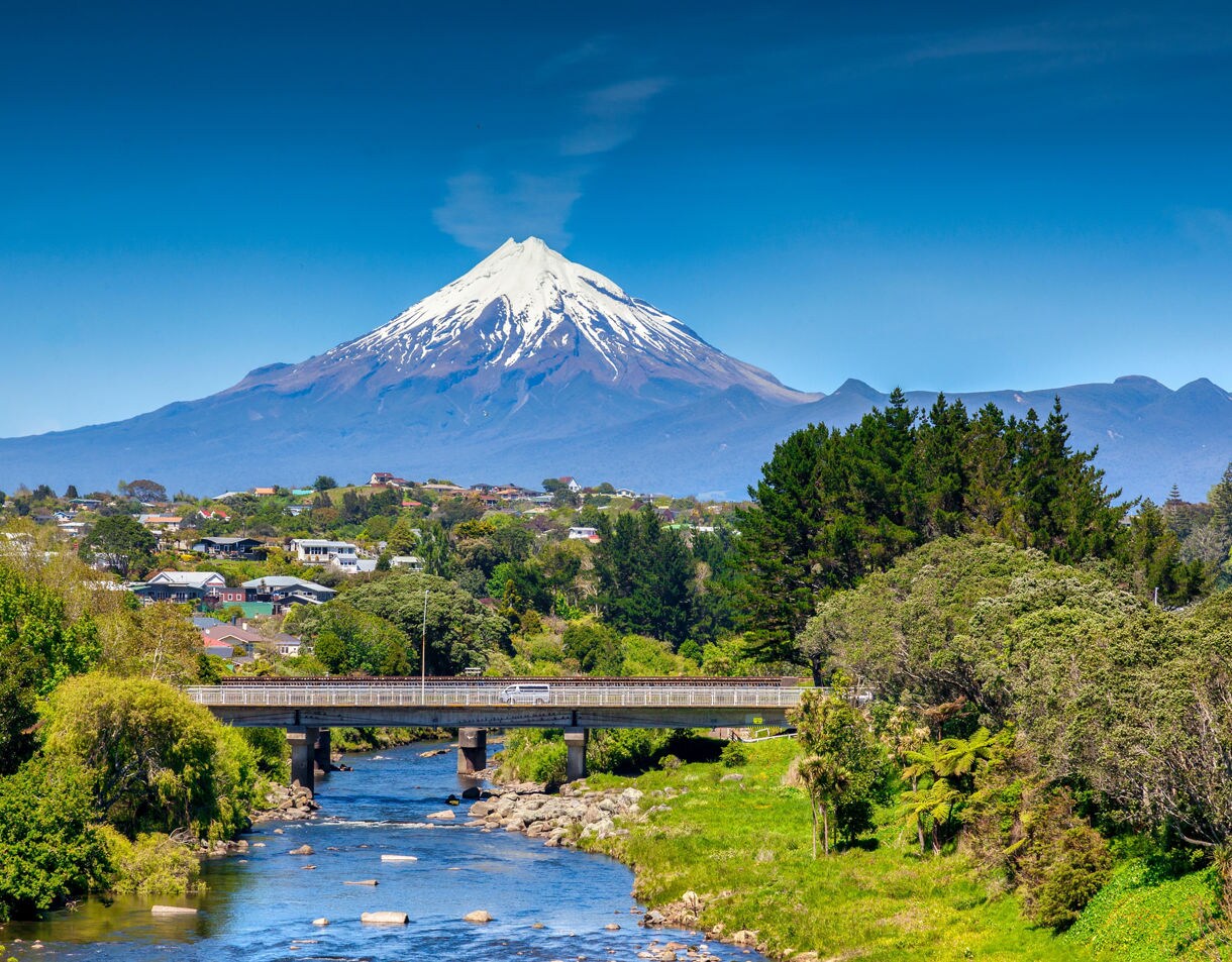 Snow-capped Mount Taranaki towering behind a small town, with a river, bridge and lush green trees in the foreground.