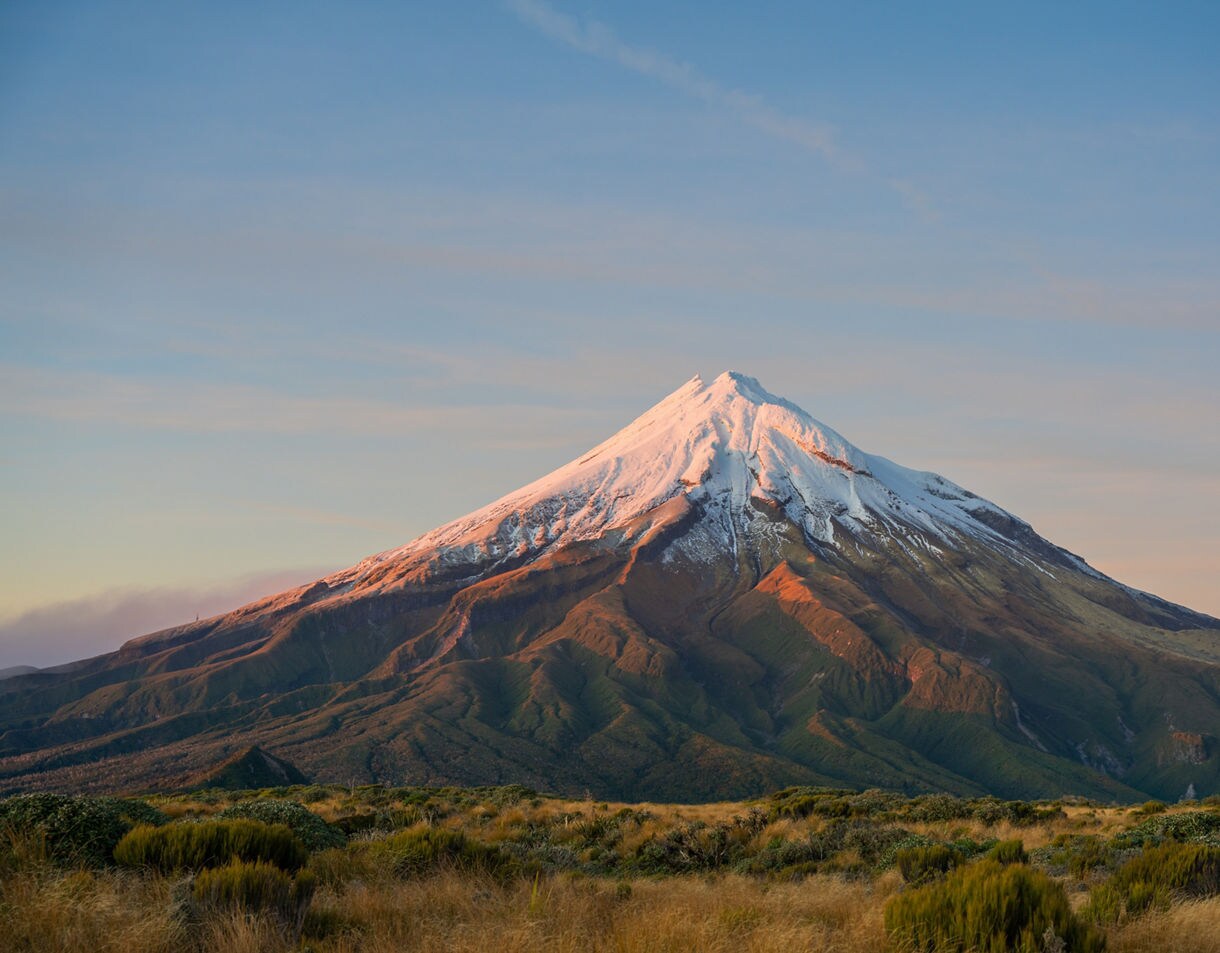 Mount Taranaki at sunrise, with its snow-covered peak glowing warm pink and golden tones above dark green lower slopes and scrubland in the foreground.