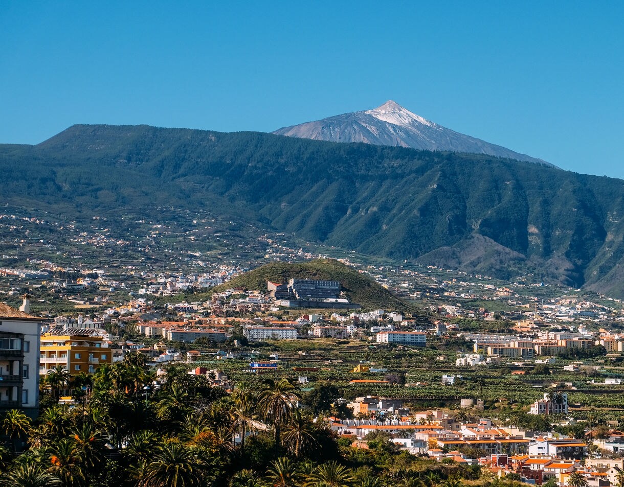 View of Mount Teide with its snow-dusted peak rising above green mountain slopes and a cityscape in Tenerife.