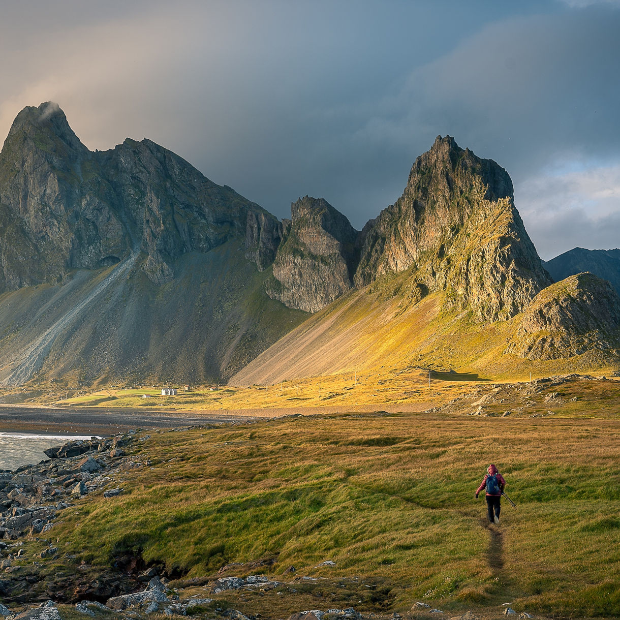 A hiker walks along a grassy path near a dark coastal shoreline, framed by dramatic, jagged volcanic mountain peaks under soft golden sunlight breaking through moody clouds.