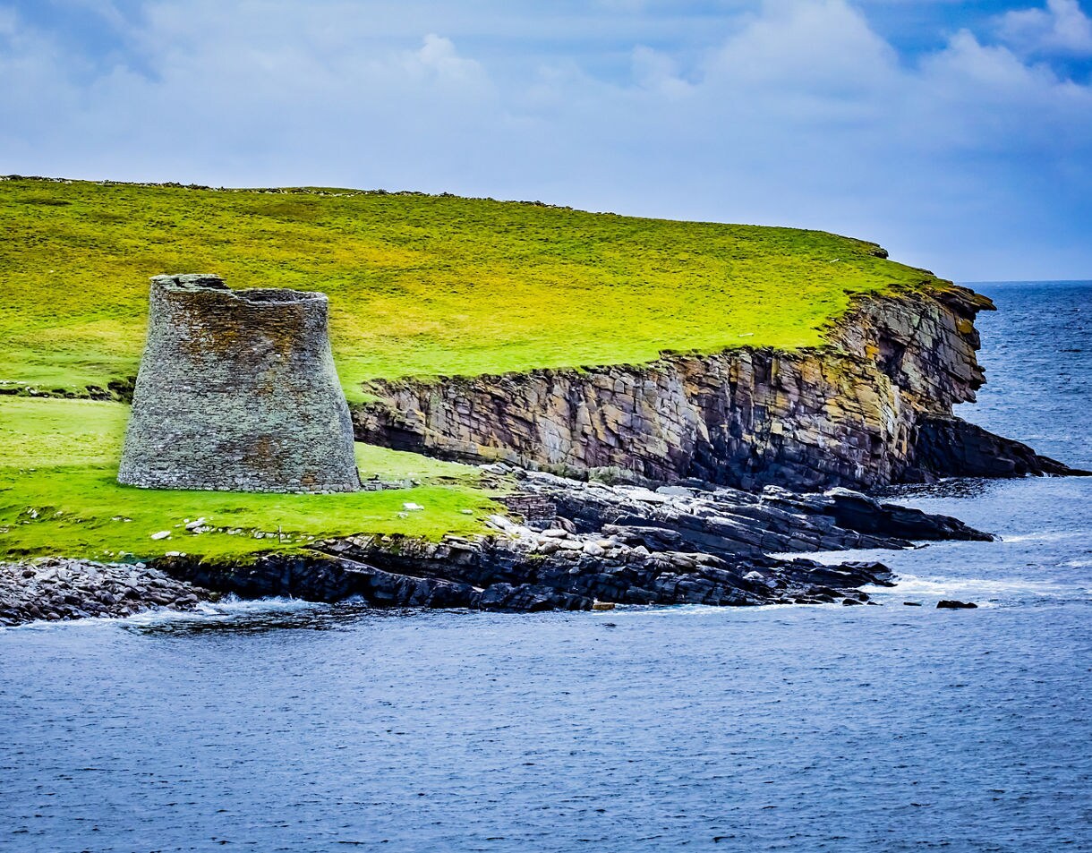 Stone broch on Mousa Island perched on green cliffs above rocky shoreline with waves crashing against the coast.
