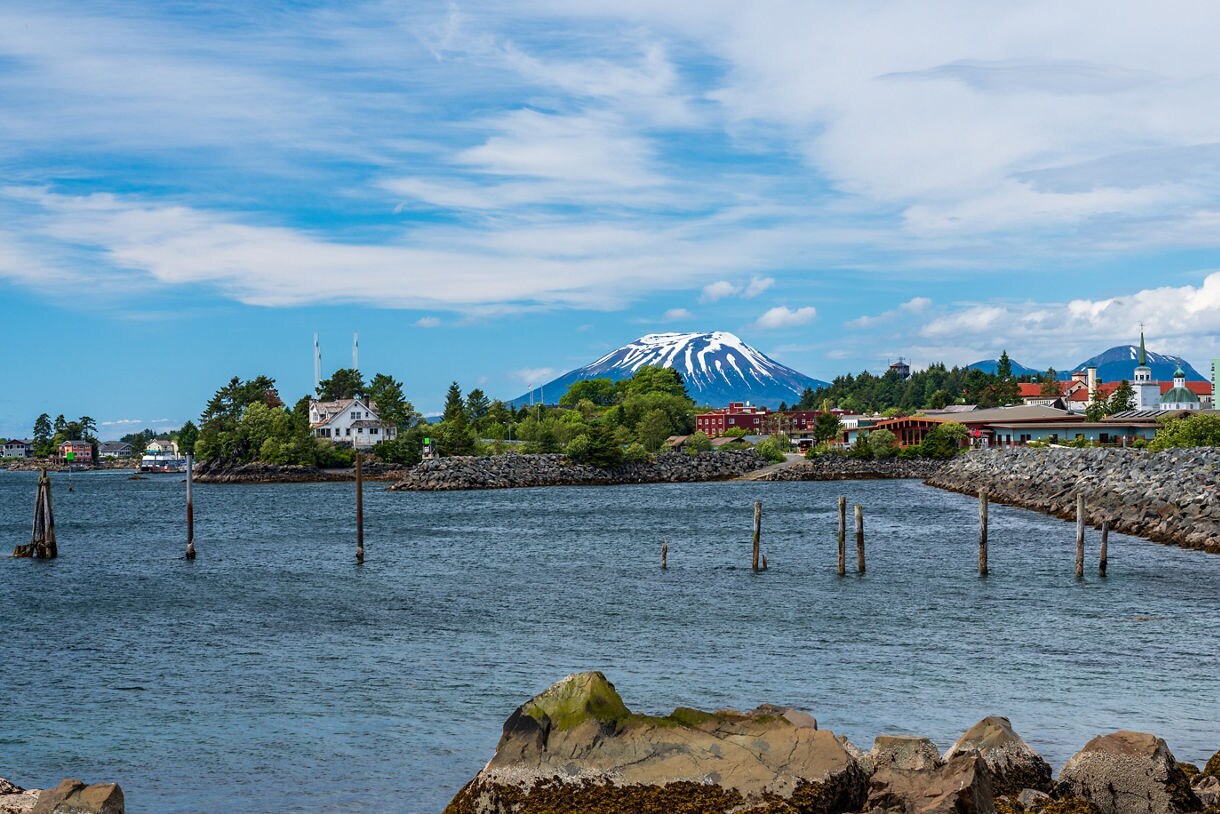 A picturesque view of Mount Edgecumbe, an extinct volcano with a snow-covered peak, towering over the harbor town of Sitka, Alaska, surrounded by rocky shores and calm waters.