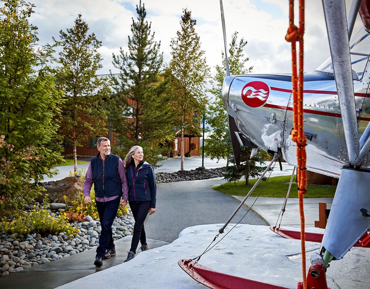 Couple walking along landscaped pathway past a large vintage seaplane sculpture with Princess Cruises logo, with lodge buildings and evergreen trees in background.