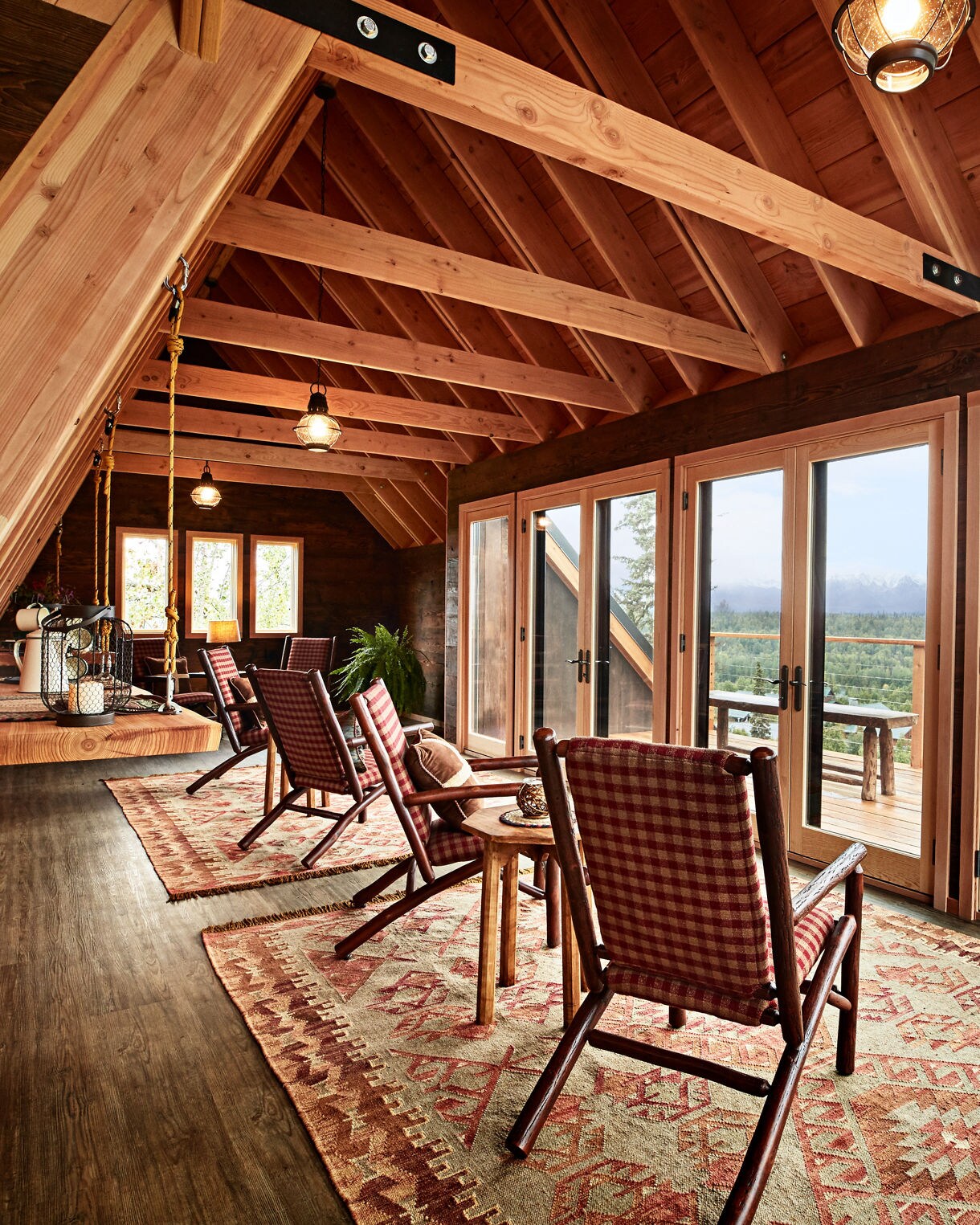Spacious lodge lounge with exposed timber beam ceiling, red buffalo plaid rocking chairs arranged on patterned rugs, pendant lighting, and French doors opening to outdoor deck with mountain views.