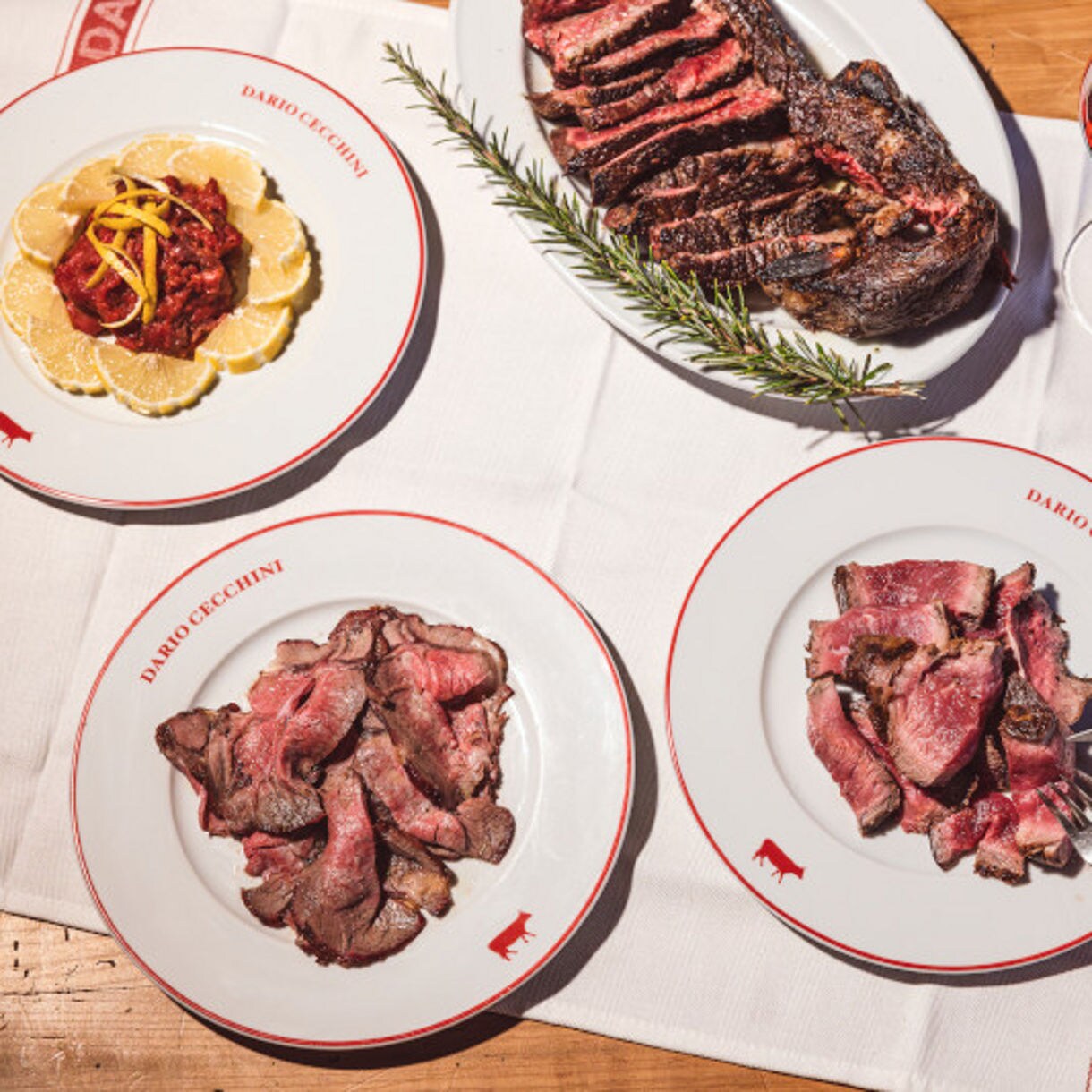 Multiple plates of sliced steaks and accompaniments arranged on a wooden table with wine glasses.