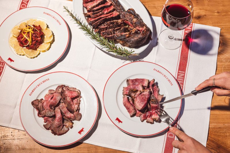 Multiple plates of sliced steaks and accompaniments arranged on a wooden table with wine glasses.