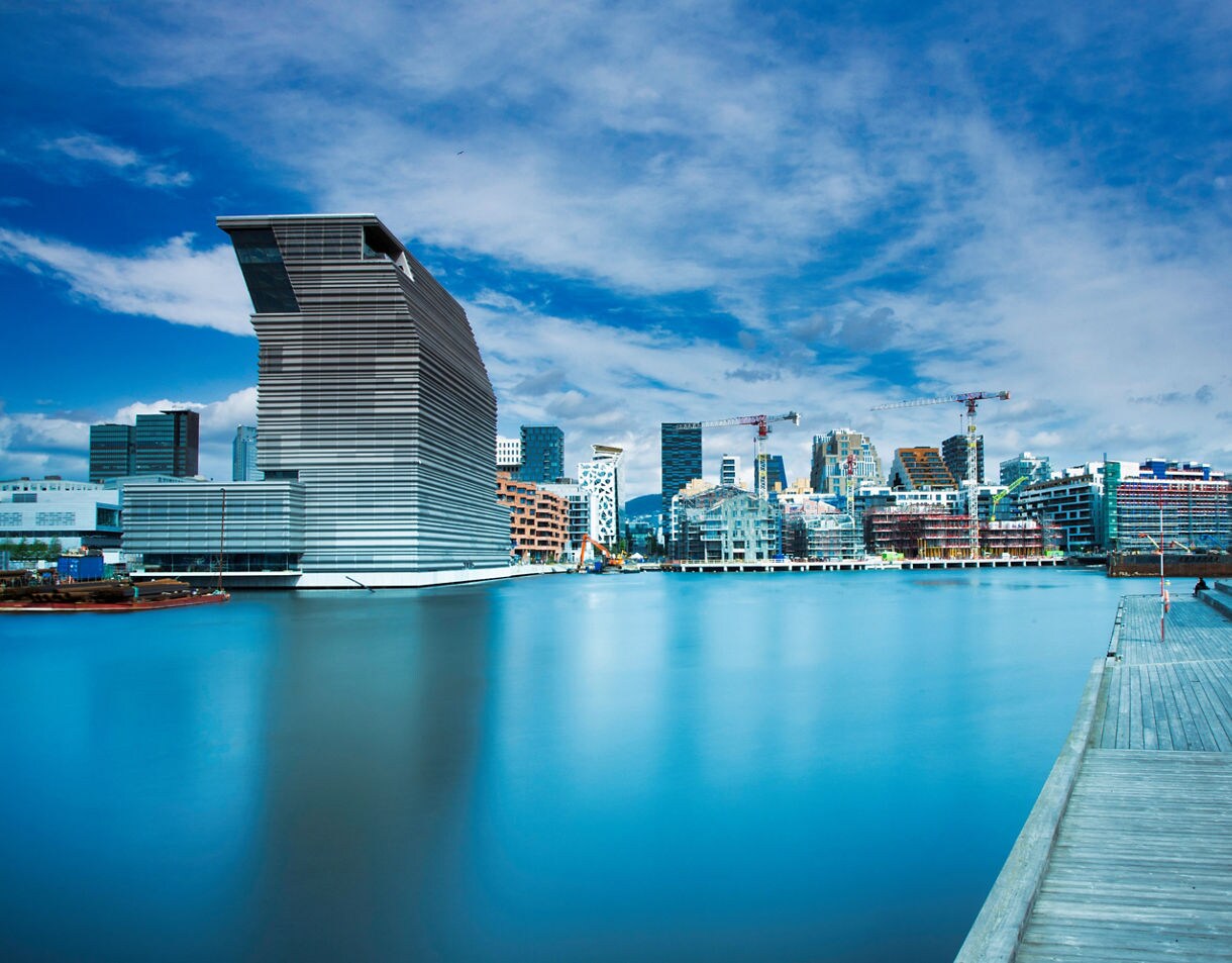 Waterfront view of the MUNCH Museum in Oslo, a modern angular glass building beside the Barcode district skyline under a partly cloudy blue sky.