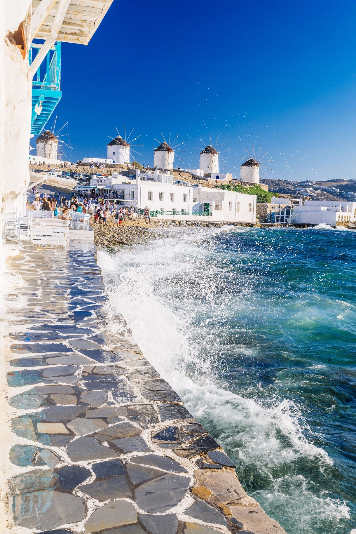 Splashing seawater along a stone walkway beside whitewashed buildings, with several historic windmills on a hill in the background.