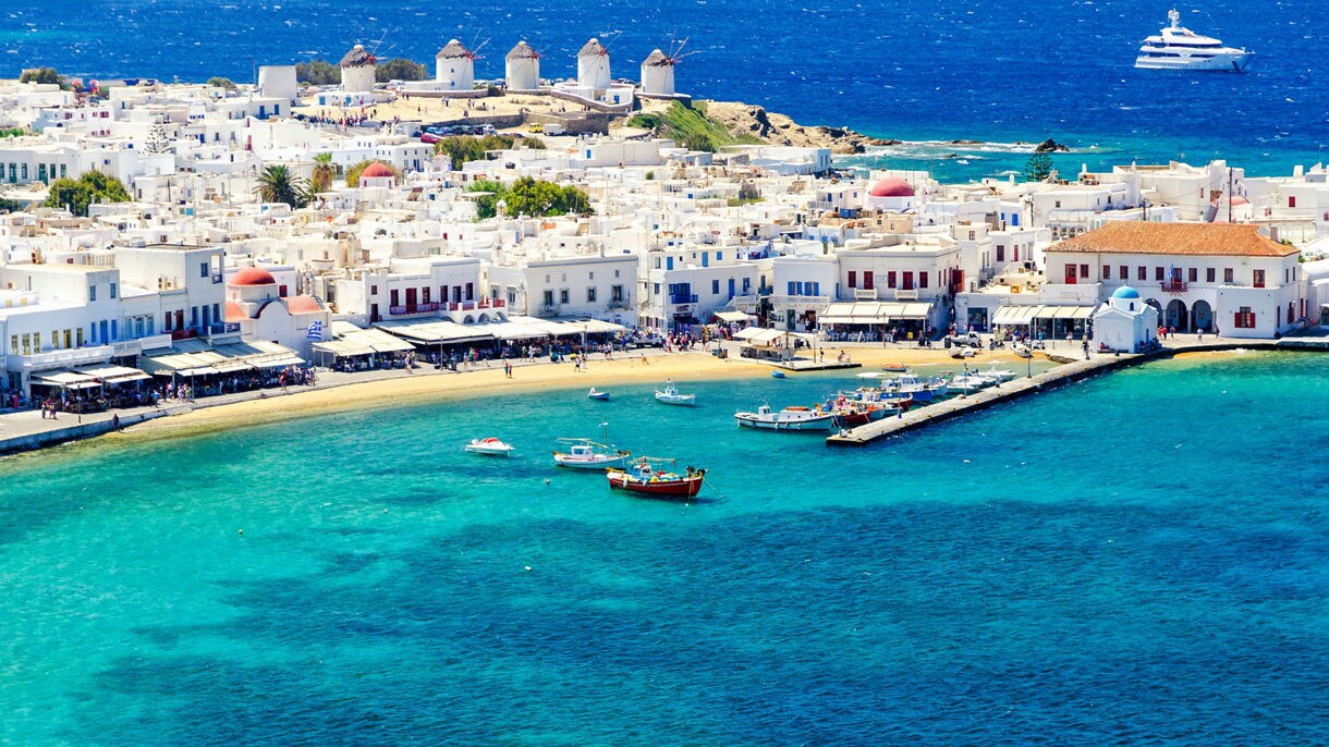 A scenic view of Mykonos, Greece, featuring traditional white buildings along the waterfront, colorful fishing boats in the clear blue water, and the island’s famous windmills in the background.