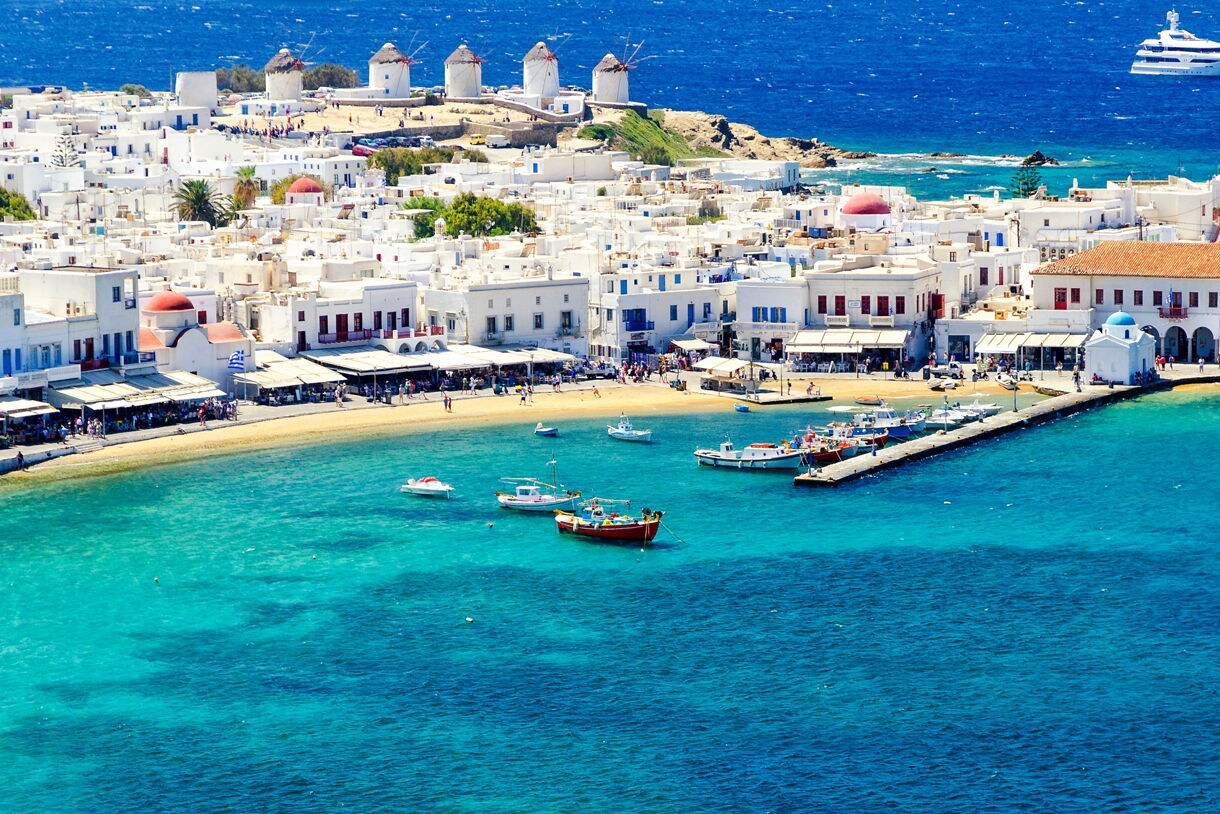 A scenic view of Mykonos, Greece, featuring traditional white buildings along the waterfront, colorful fishing boats in the clear blue water, and the island’s famous windmills in the background.