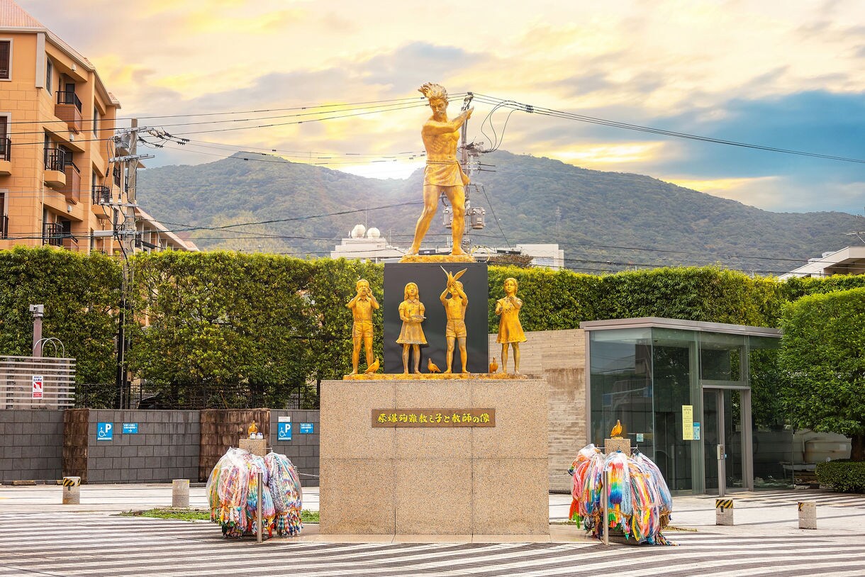 Golden statues of children and a man stand on a stone pedestal at the Nagasaki Atomic Bomb Museum in Japan, with colorful paper cranes laid in tribute and green hedges, mountains and buildings visible in the background under a glowing sky.