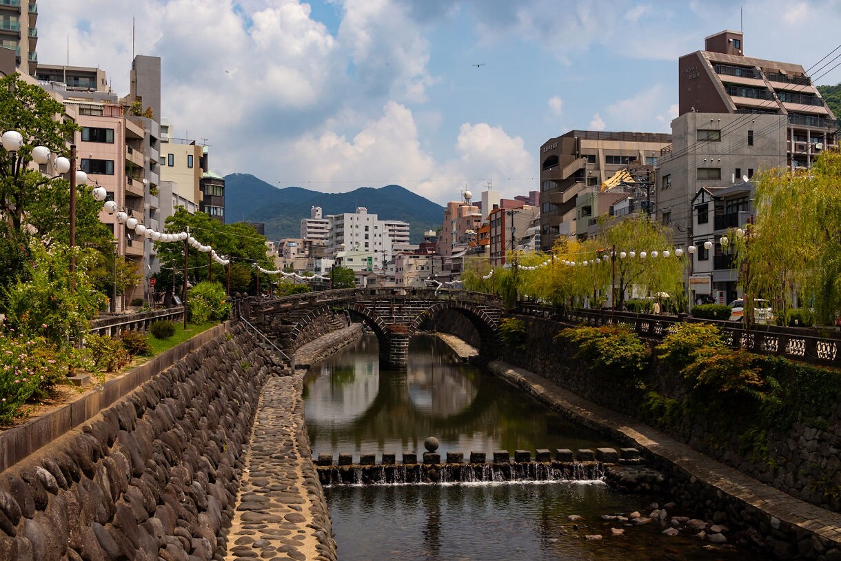 View of the Megane Bridge in Nagasaki, Japan, spanning a calm canal lined with stone walls, lanterns, trees and urban buildings under a partly cloudy sky with mountains in the distance.