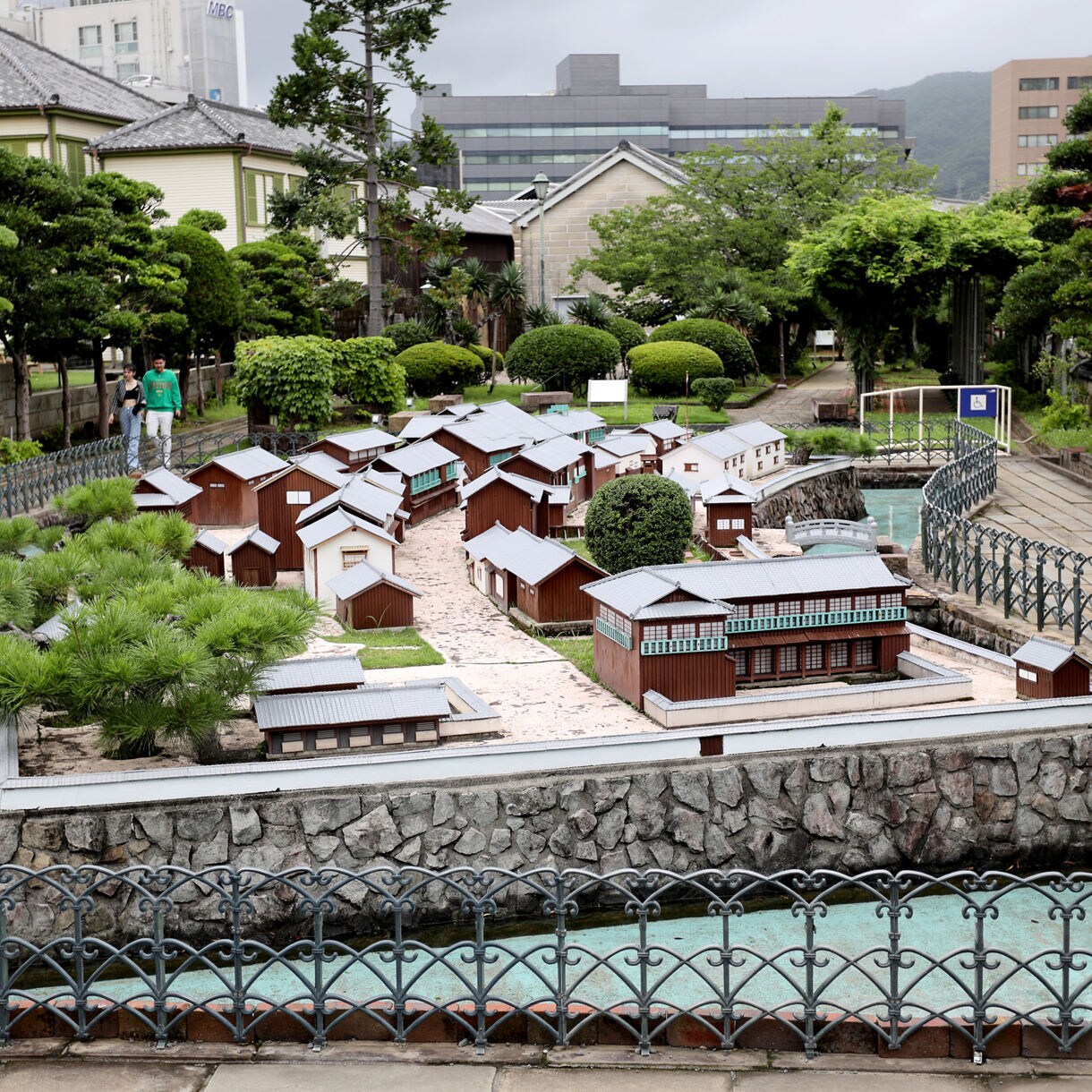 A scale model of Dejima, the historic Dutch trading post in Nagasaki, Japan, featuring rows of miniature wooden buildings, stone walls and water canals, surrounded by manicured greenery and modern city buildings in the background.