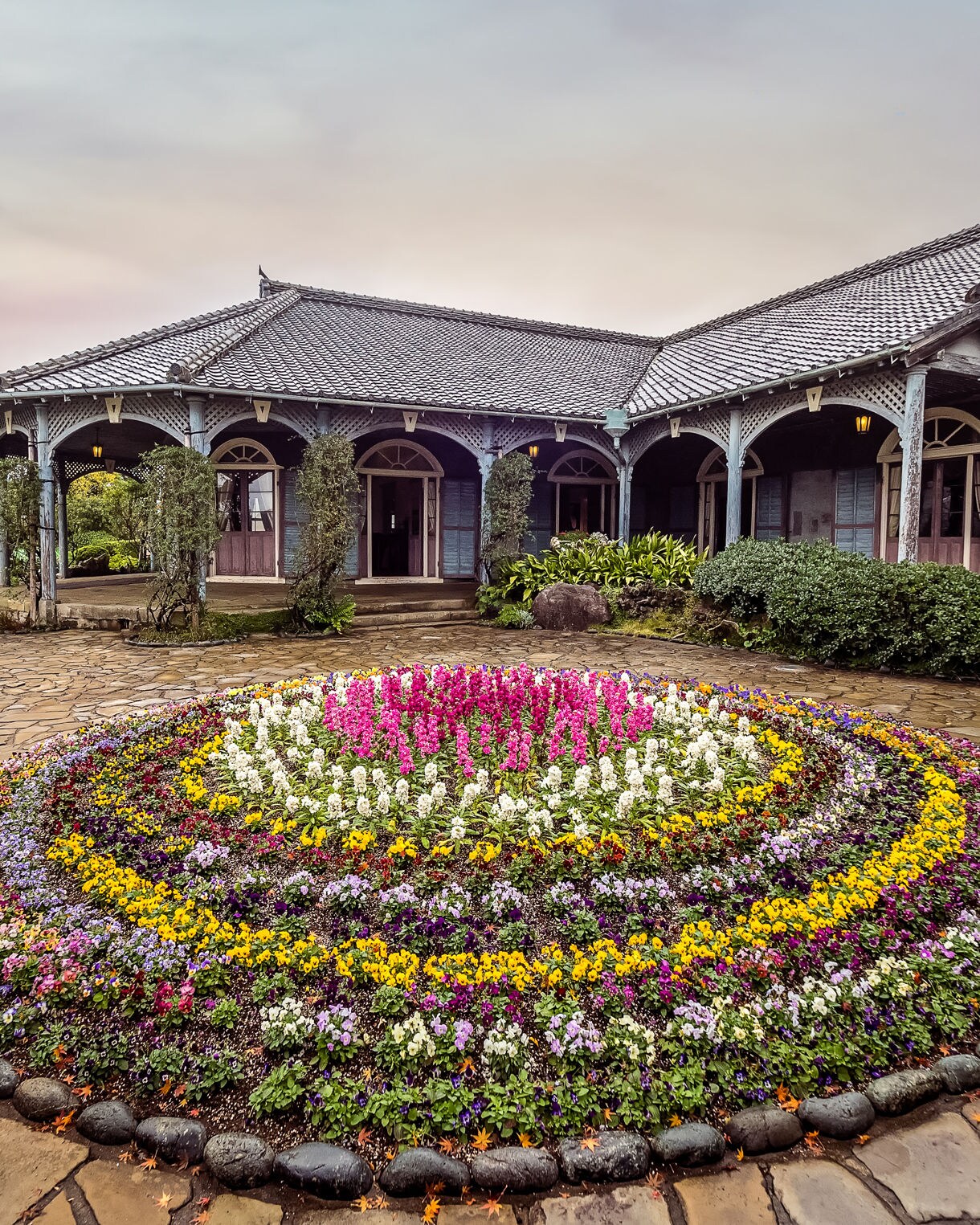A circular flower bed bursting with colorful blooms sits in front of a traditional Western-style residence with arched verandas and tiled rooftops at Glover Garden in Nagasaki, Japan. The stone path and lush greenery surround the historic home under a soft, overcast sky.