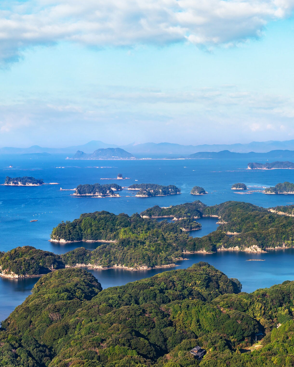 Aerial view of the Kujūku Islands in Nagasaki, Japan, showing dozens of small, forest-covered islands scattered across a vibrant blue ocean under a partly cloudy sky, with distant mountains on the horizon.