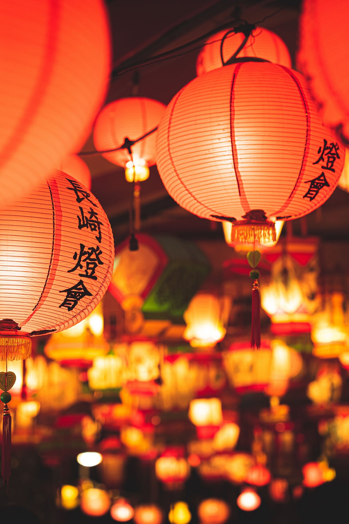 Close-up of red and orange paper lanterns with Chinese characters glowing warmly during the Nagasaki Lantern Festival, with a blurred background filled with colorful hanging lights creating a festive atmosphere.