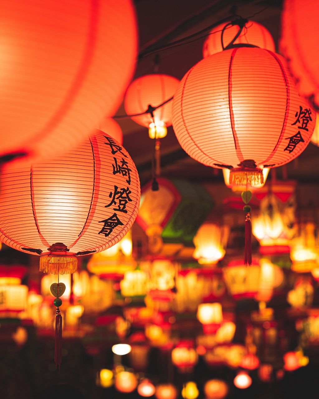 Close-up of red and orange paper lanterns with Chinese characters glowing warmly during the Nagasaki Lantern Festival, with a blurred background filled with colorful hanging lights creating a festive atmosphere.