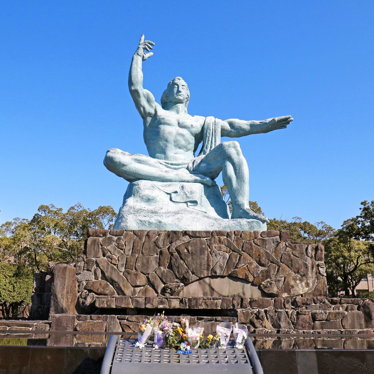 The Peace Statue at Nagasaki Peace Park in Japan, a large bronze figure seated with one arm raised and one extended, set atop a stone pedestal with flowers laid in remembrance, surrounded by trees.