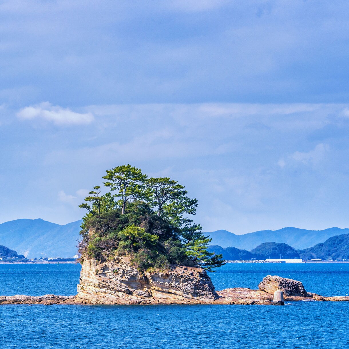 Small rocky island covered with green pine trees in the middle of a calm blue sea.