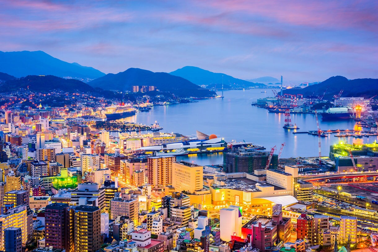 Panoramic view of Nagasaki, Japan at dusk, showing a brightly lit cityscape with colorful buildings, a harbor filled with cruise ships and cranes and surrounding mountains under a vibrant pink and blue sky.