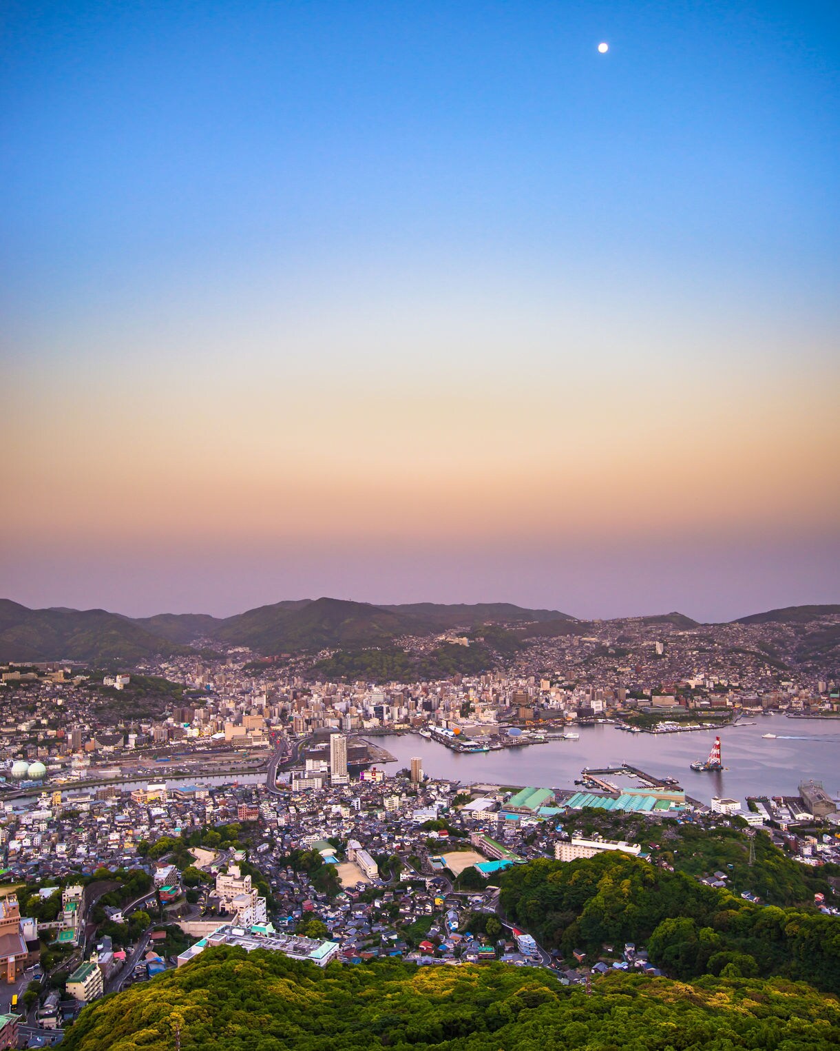 A twilight aerial view of Nagasaki, Japan showing the city’s urban sprawl and harbor nestled between forested hills, with a pastel sky fading from orange to deep blue and a bright moon visible overhead.