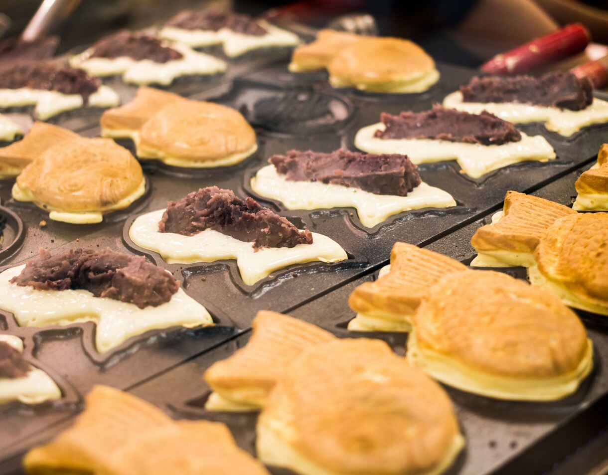  Fish-shaped taiyaki pastries being cooked on a griddle, filled with red bean paste in various stages of preparation