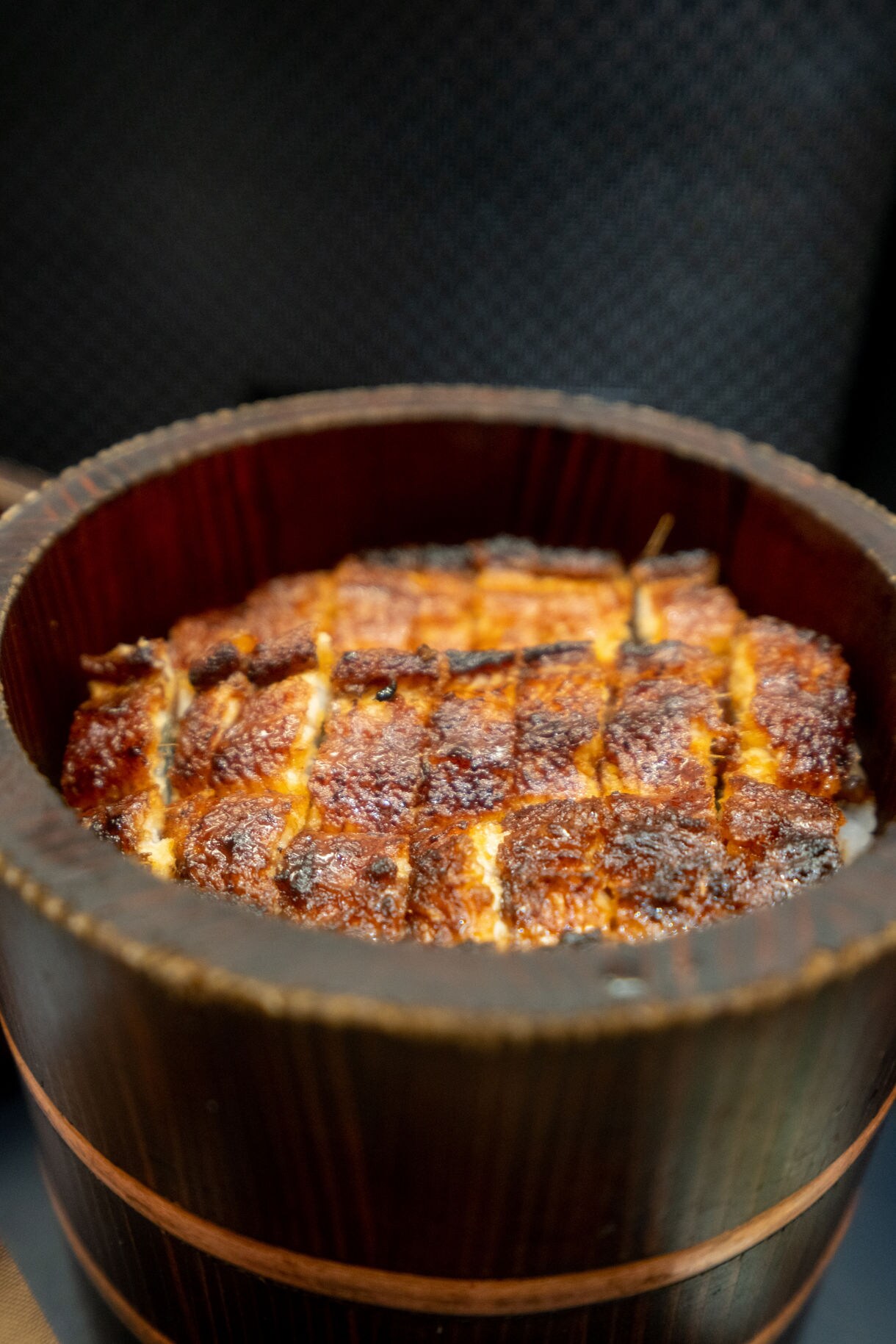 Close-up of sliced grilled eel glazed with a glossy sauce, served over rice in a traditional wooden bowl.