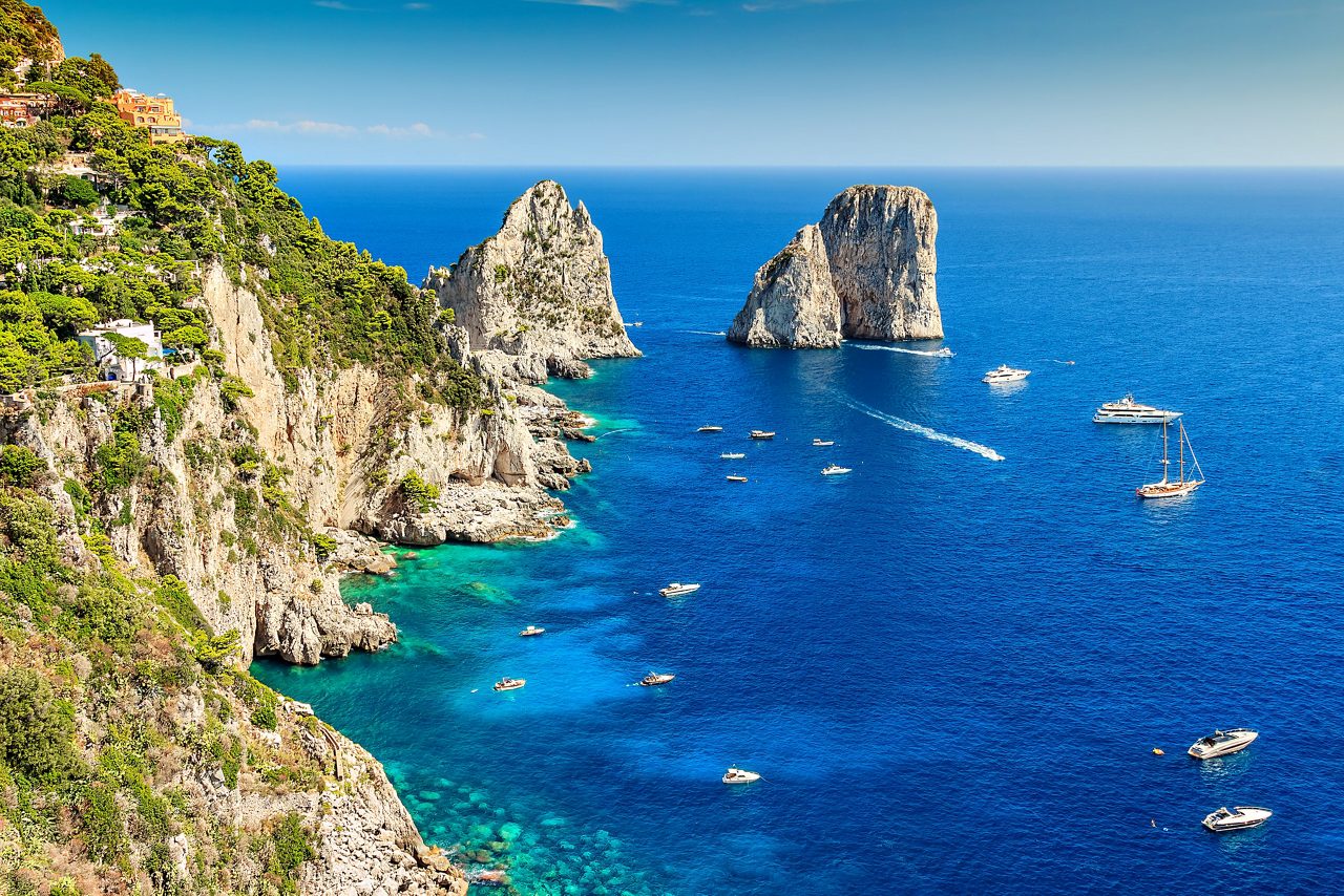 Steep cliffs of Capri with turquoise waters and boats circling the famous Faraglioni rock formations.