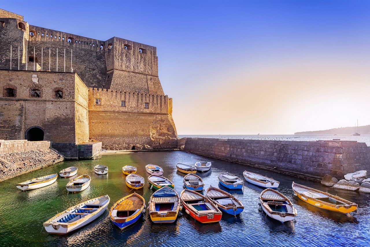 Rowboats floating in the harbor beside Castel dell’Ovo, a medieval seaside fortress in Naples, at sunset.