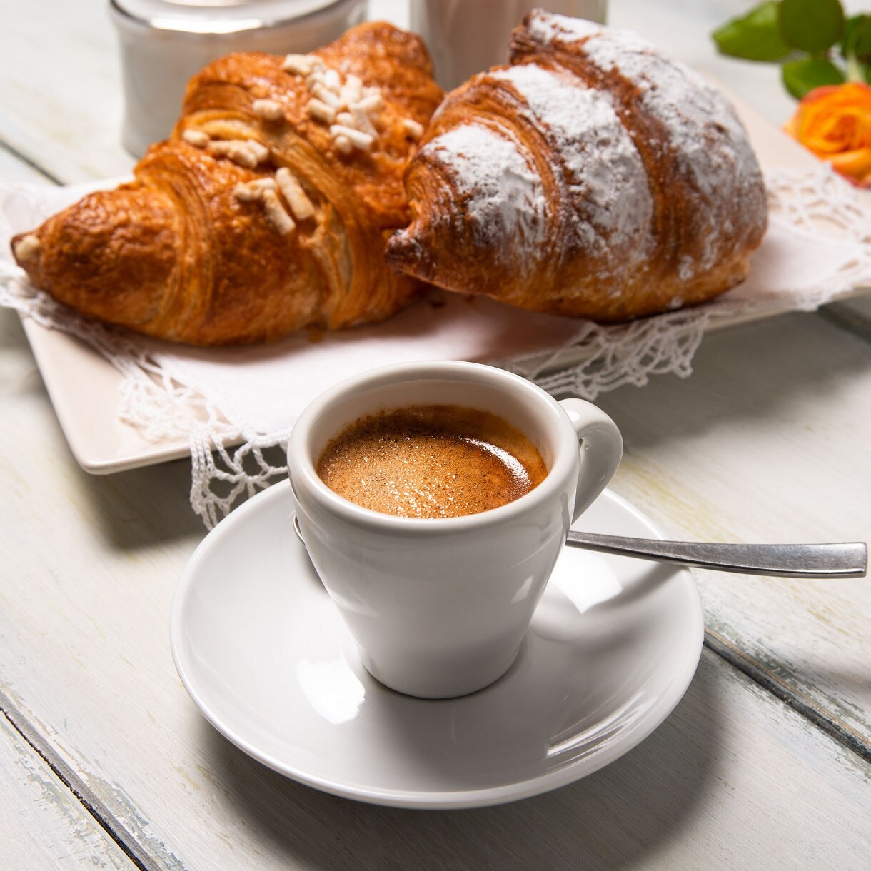 Close-up of a cup of espresso with two croissants dusted with sugar and nuts on a plate in the background.