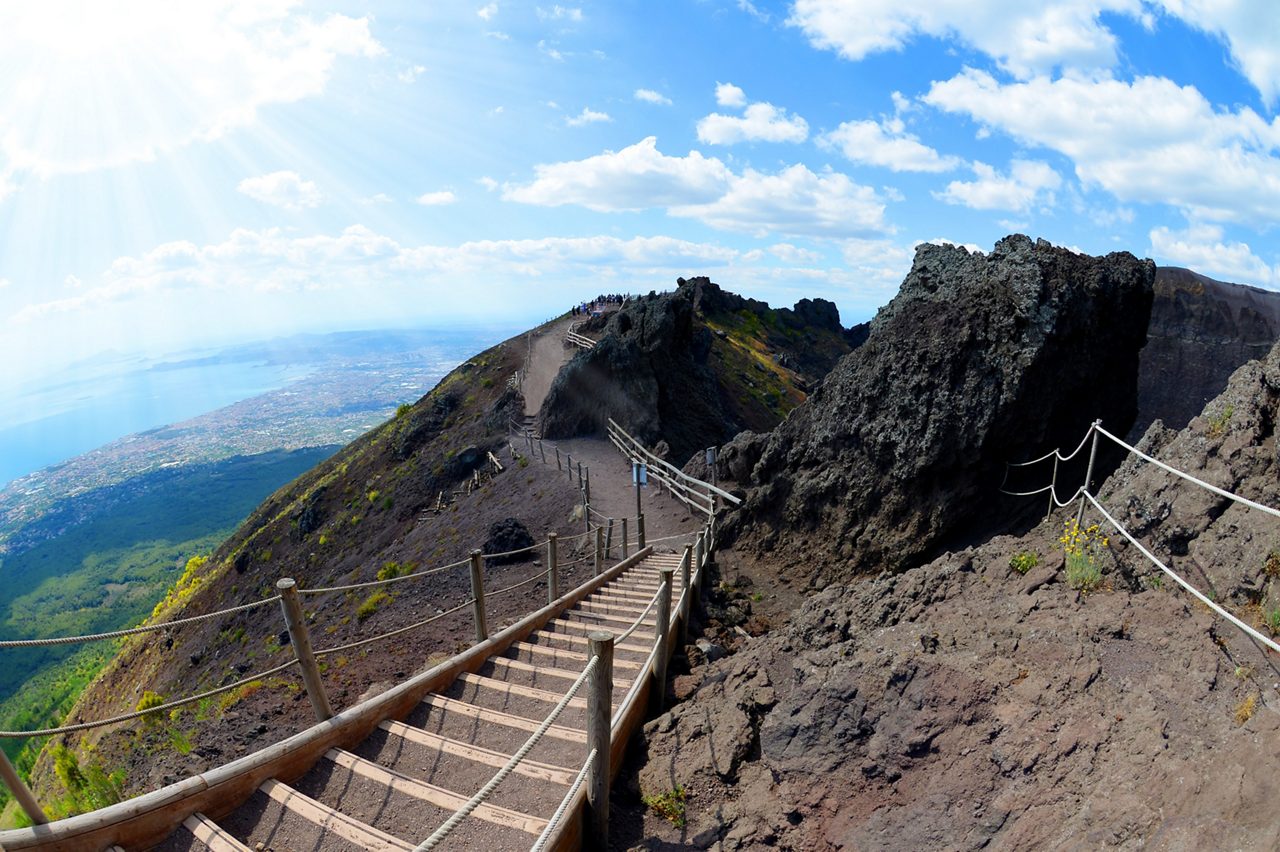 Wooden stairway winds up Mount Vesuvius with rocky slopes and a panoramic view of the coast below.