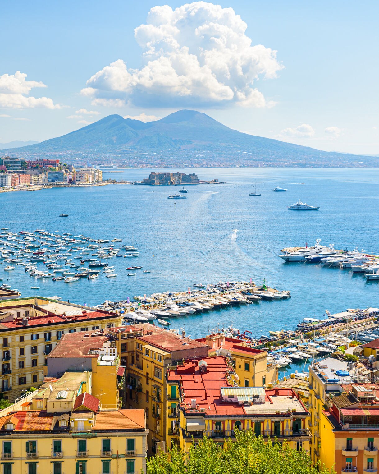 View of Naples waterfront with colorful buildings, harbors filled with boats and Mount Vesuvius in the background.
