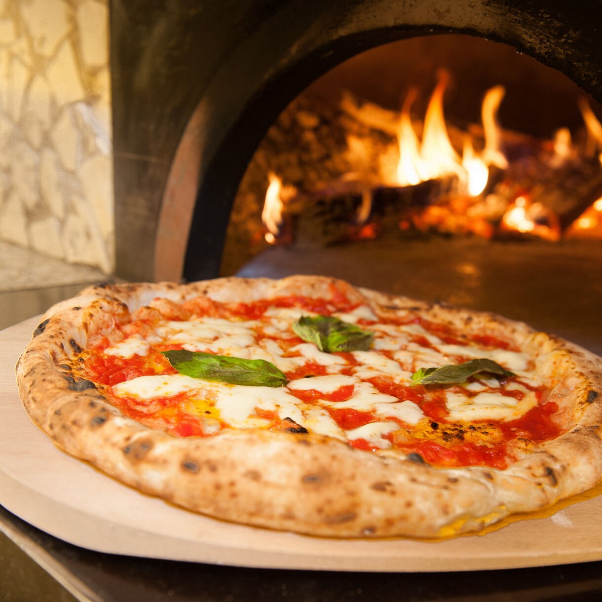 A Neapolitan-style Margherita pizza with charred crust, fresh basil leaves, cherry tomatoes, and mozzarella cheese displayed on a red and white checkered tablecloth.