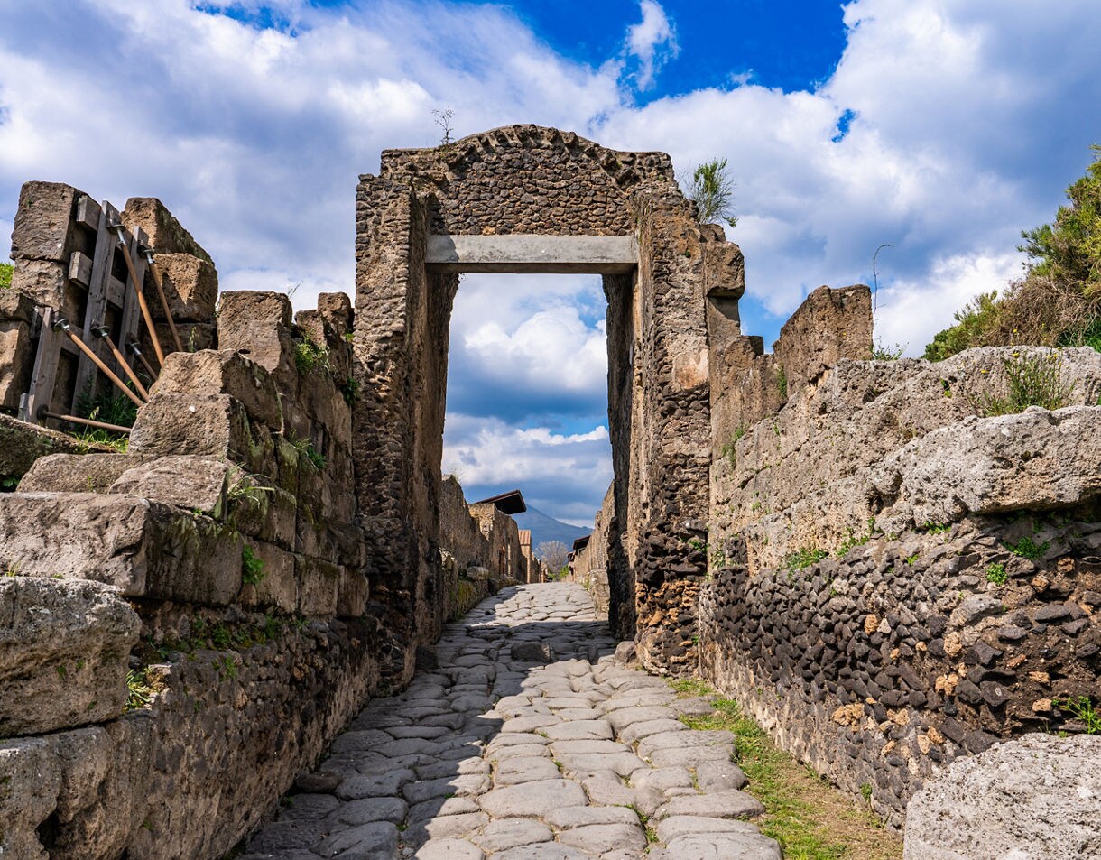 Stone archway and cobbled street among the ruins of Pompeii under a partly cloudy sky.