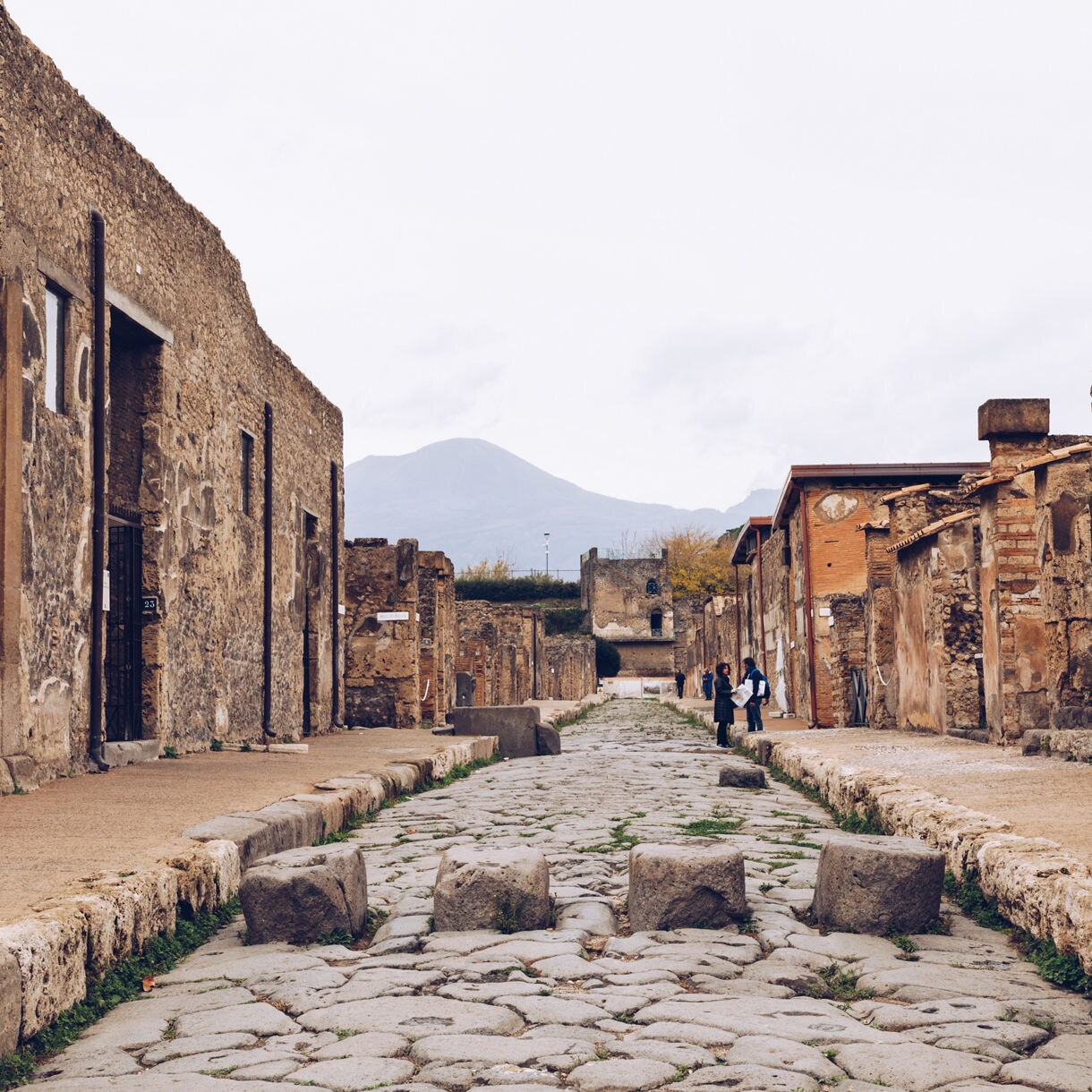 Cobblestone street lined with ruins in Pompeii, with Mount Vesuvius visible in the background.