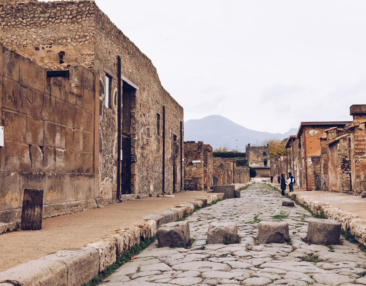 Stone-paved street lined with ruins in Pompeii, with Mount Vesuvius visible in the background.
