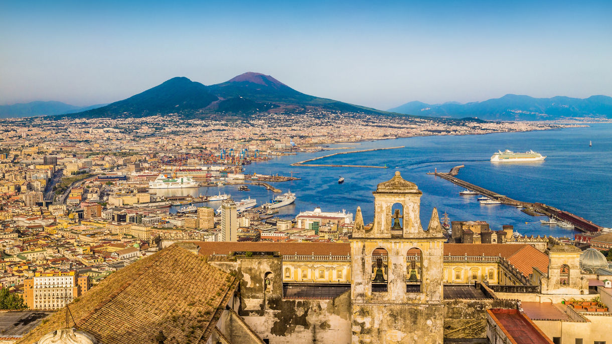 Aerial view of Naples with a historic bell tower in the foreground, a busy harbor with cruise ships and Mount Vesuvius rising in the background.