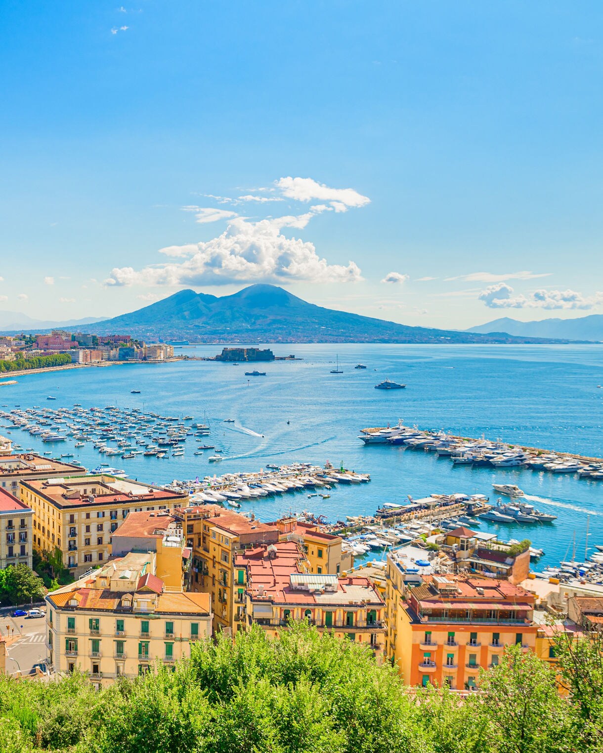 Panoramic view of Naples with colorful buildings, a marina filled with boats and Mount Vesuvius in the distance.