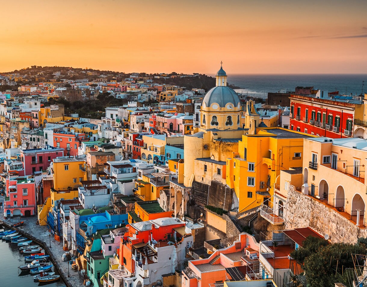 Colorful buildings and boats in Procida, Italy, glowing under a golden sunset by the sea.