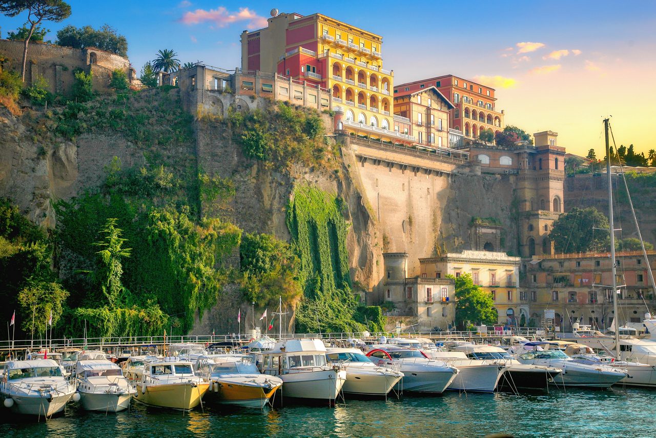 Boats docked in a marina below towering cliffs, topped with colorful villas in Sorrento at sunset.