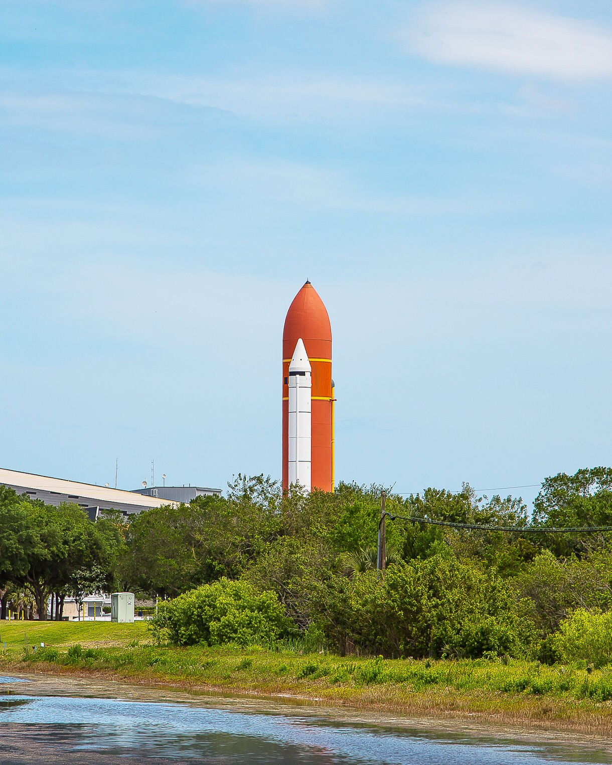 NASA's Kennedy Space Center in Cape Canaveral, Florida, featuring a large orange and white rocket standing upright in a field, surrounded by trees and a body of water, with a building in the background under a blue, cloudy sky.