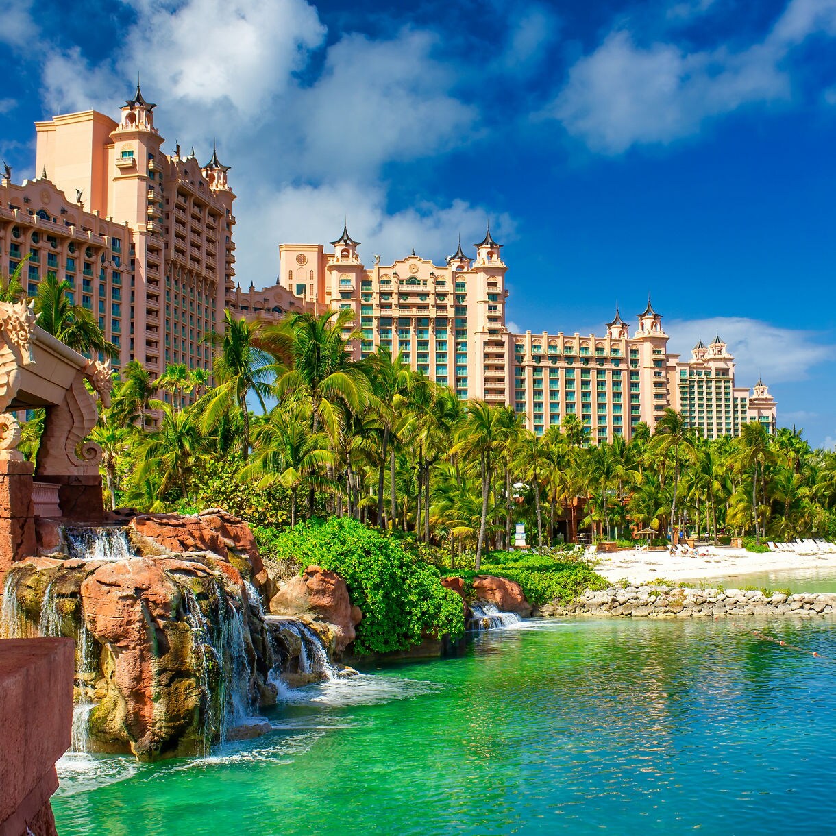 View of the Atlantis Resort in Nassau with pink towers, palm trees, waterfalls and a white sand beach beside clear turquoise water under a bright blue sky.