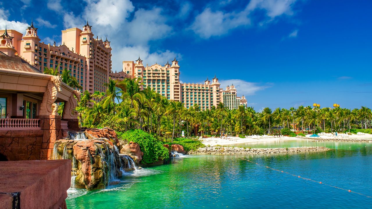View of the Atlantis Resort in Nassau with pink towers, palm trees, waterfalls and a white sand beach beside clear turquoise water under a bright blue sky.