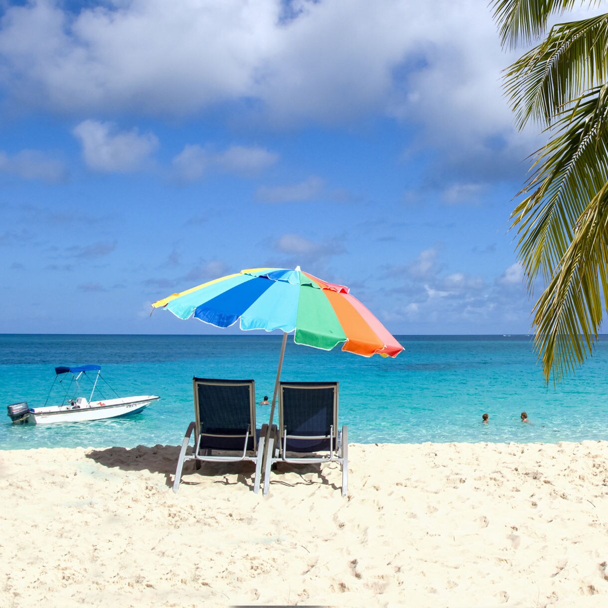 Two beach chairs under a multicolored umbrella on soft white sand beside turquoise water, with palm fronds and a small boat in the background on Blue Lagoon Island.