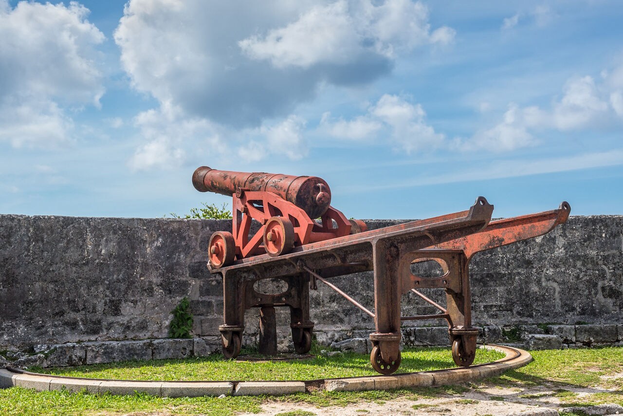 A weathered red cannon mounted on an iron carriage at Fort Fincastle in Nassau, set against a stone wall with a bright blue sky and scattered clouds in the background.