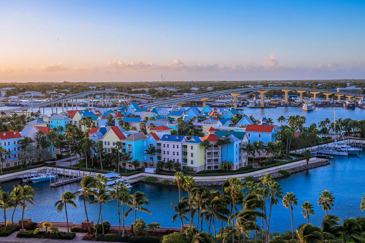 Aerial view of colorful resort buildings with red, green and blue roofs on the waterfront in Nassau, with palm trees, yachts and a bridge in the background.