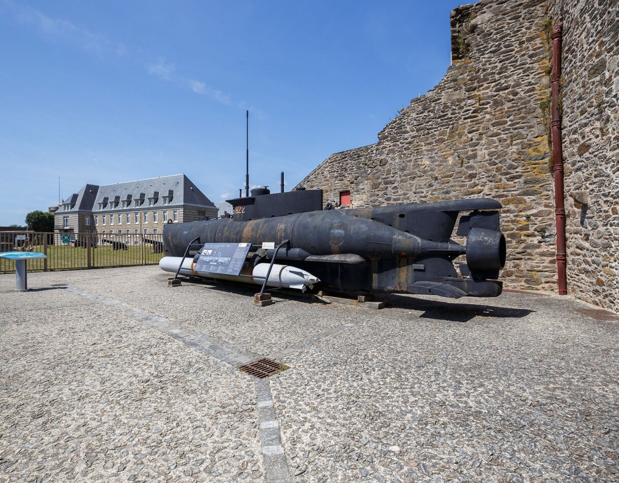 A small, weathered World War II submarine sits outdoors against stone ramparts, with a museum building and clear blue sky in the background.