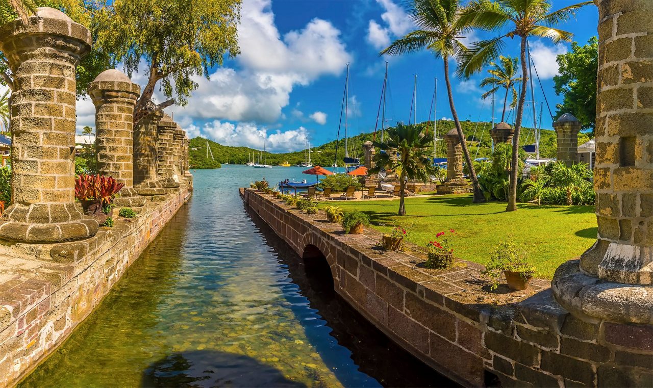 Stone archway and colonial buildings of historic Nelson's Dockyard with harbor view, sailboat masts, palm trees, and manicured grounds in Antigua.